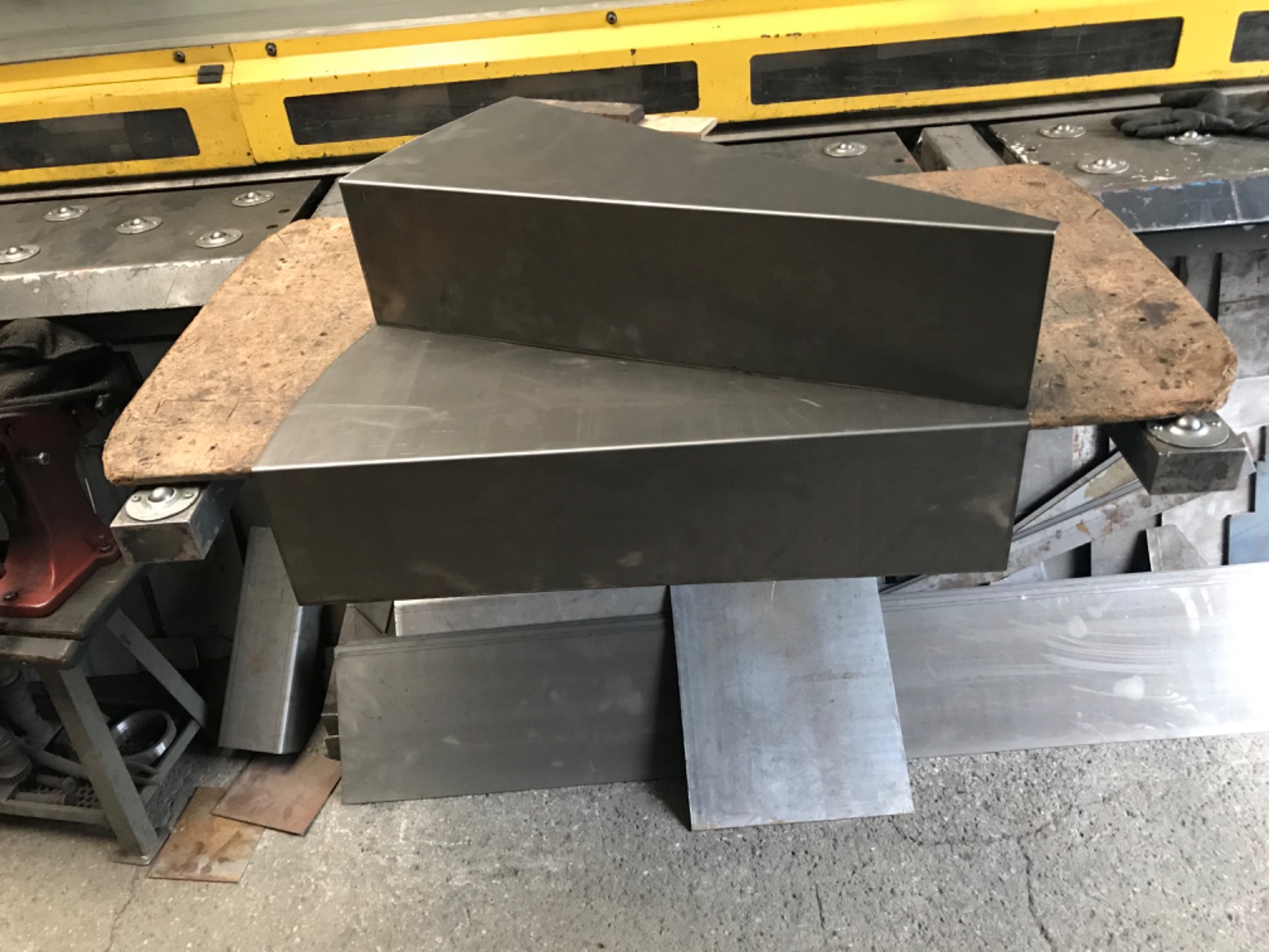 Dwa metalowe elementy o trójkątnym przekroju, ułożone jeden na drugim na warsztatowym stole, w tle widoczna maszyna do obróbki metali.