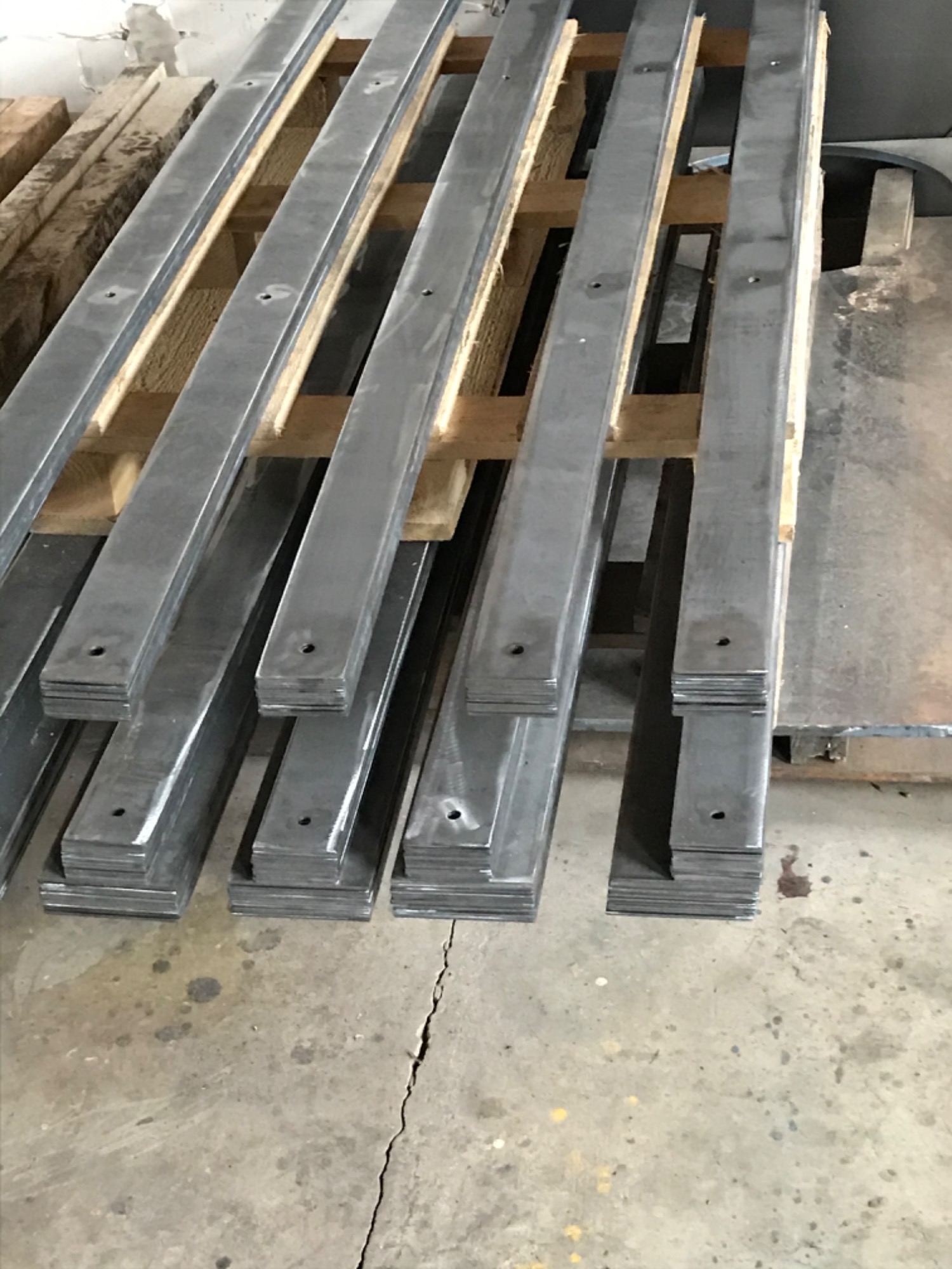 Stalowe płaskowniki z otworami ułożone na drewnianej palecie w warsztacie, widoczna surowa powierzchnia metalu i ślady obróbki.