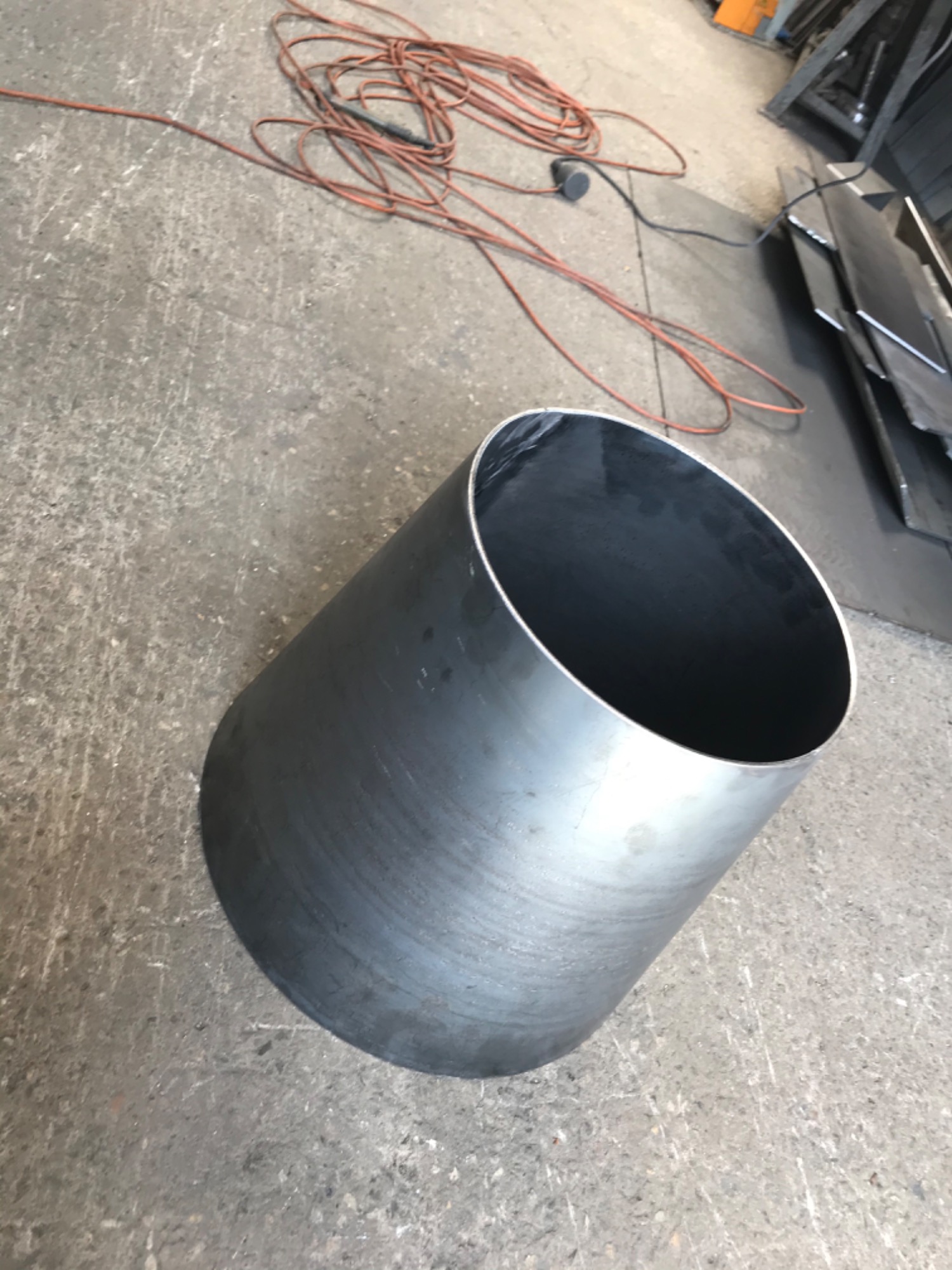 Cylindryczny element z surowej blachy stalowej, widoczny na tle betonowej posadzki warsztatu, z fragmentami kabla zasilającego i arkuszami blachy w tle.