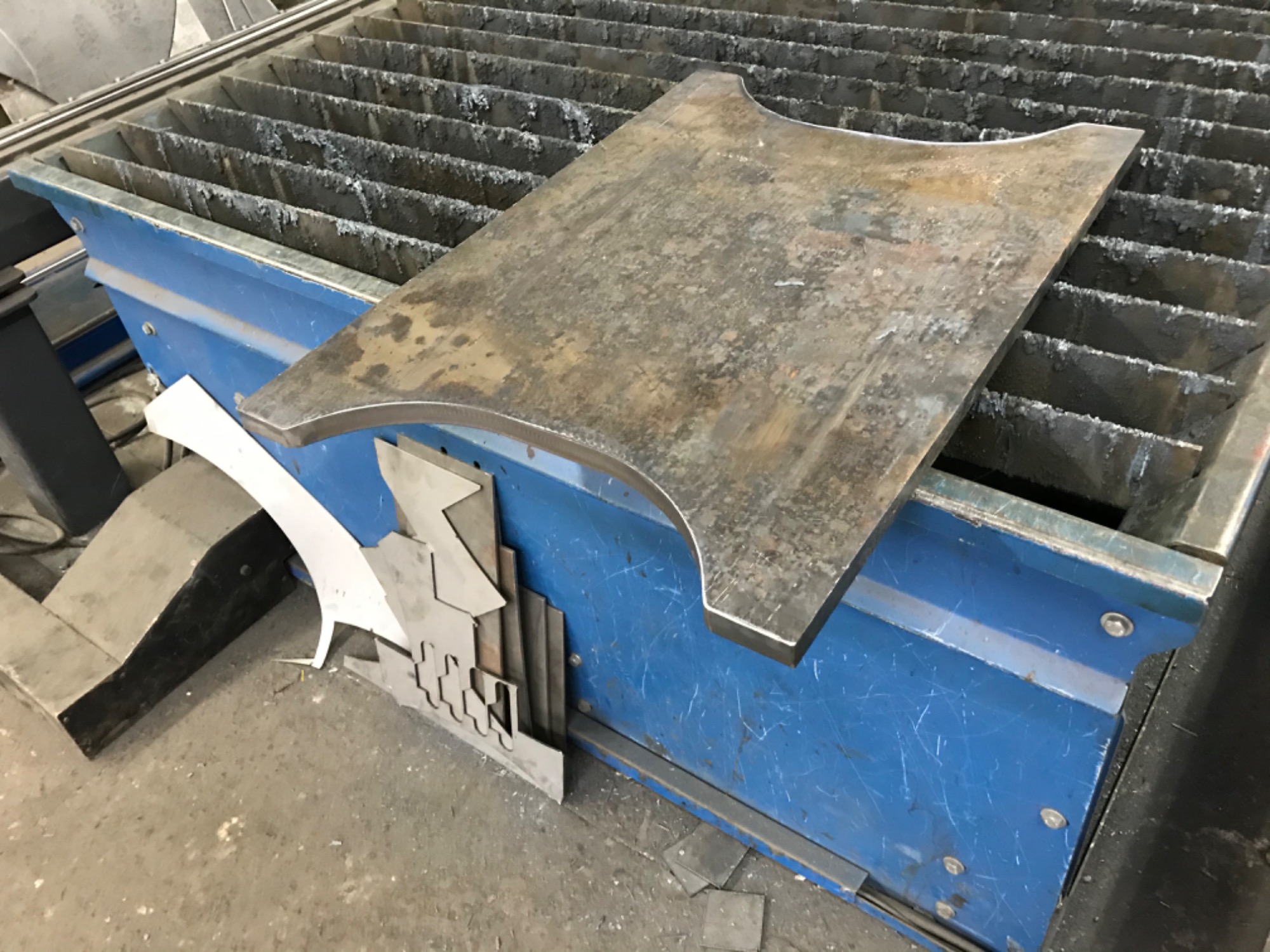 Surowy, wycięty element z blachy o nieregularnym kształcie, leżący na niebieskim stole roboczym z metalowymi listwami. Obok widoczne są inne, mniejsze fragmenty blachy i odpady po cięciu.
