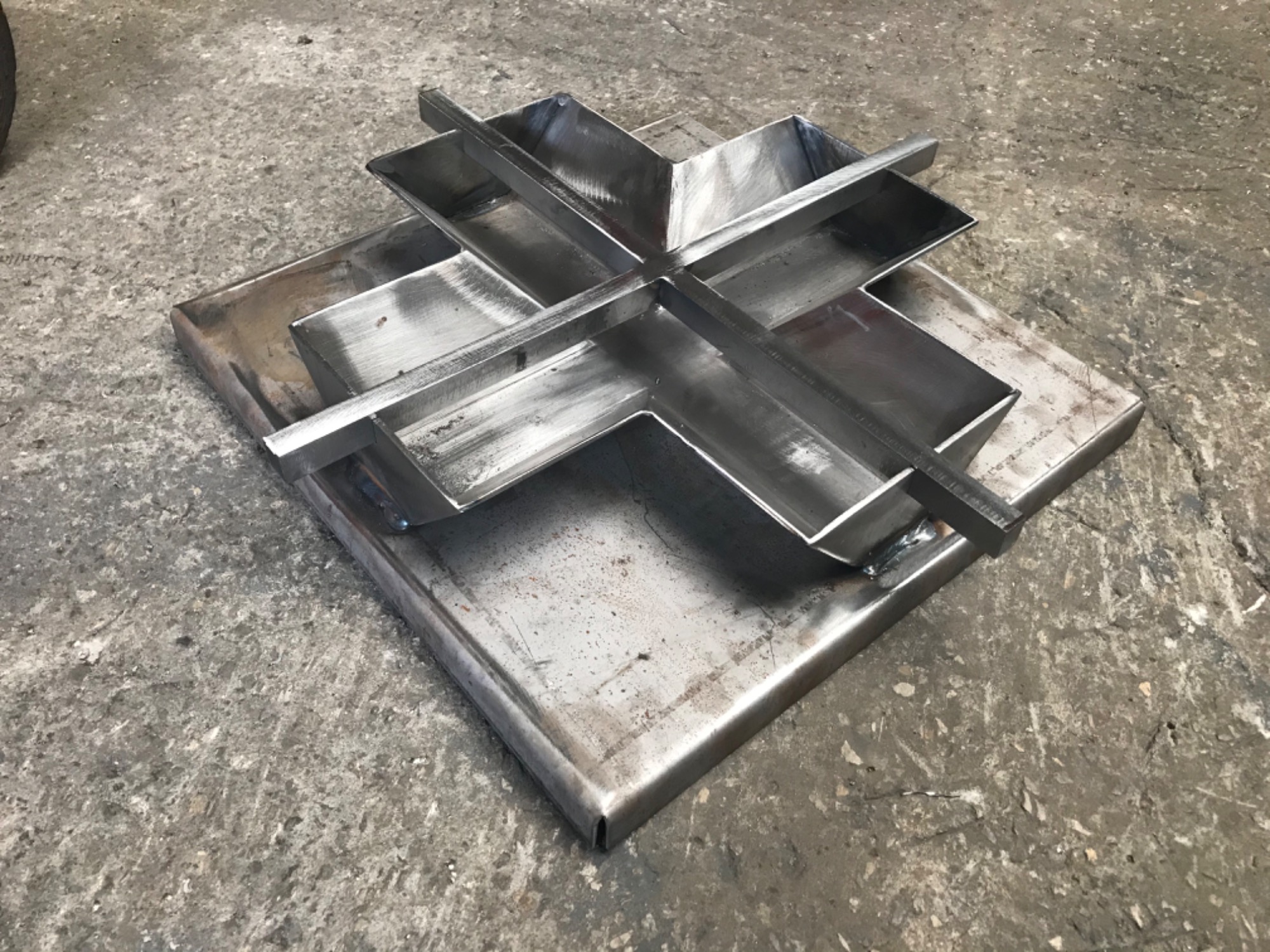 Surowa, spawana konstrukcja krzyżowa z metalowych profili na kwadratowej podstawie, widoczne spawy, szlifowane krawędzie, na tle betonowej posadzki.