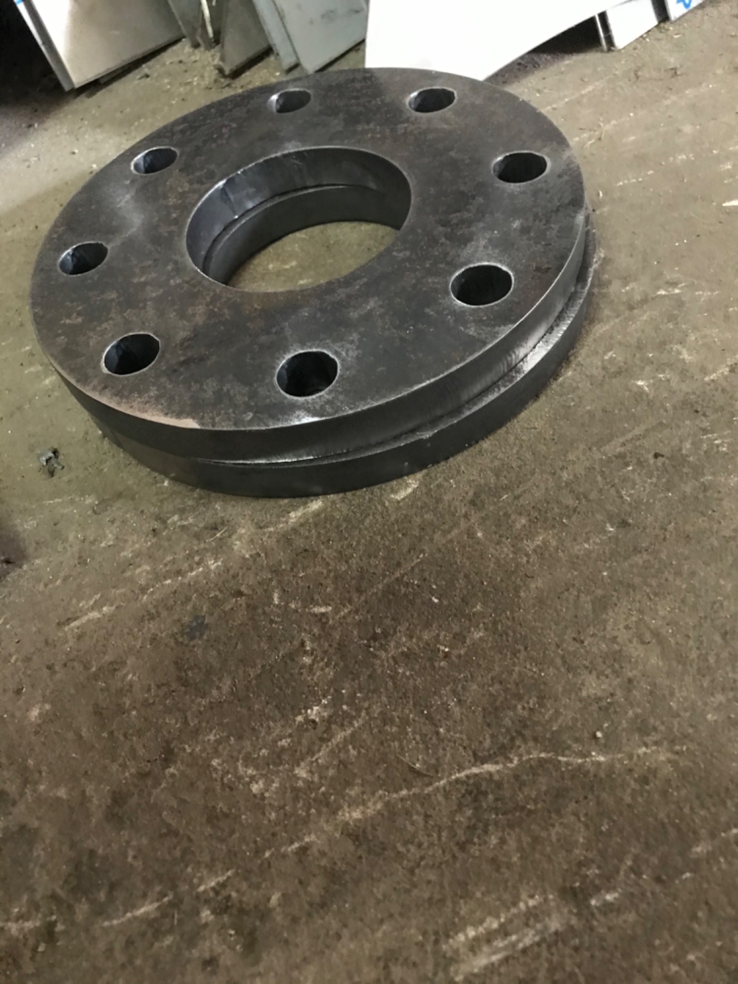 Dwie metalowe, okrągłe flansze z otworami na śruby, ułożone jedna na drugiej na betonowej podłodze. Widoczne ślady obróbki i patyna.