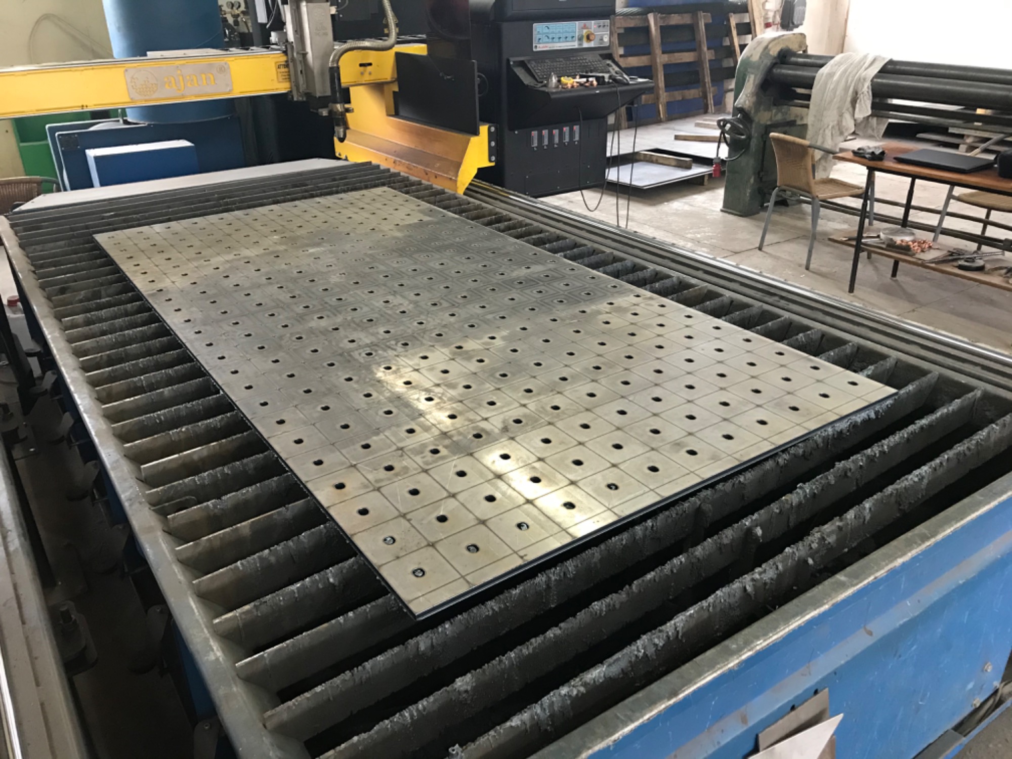 Proces cięcia blachy perforowanej na maszynie CNC w zakładzie w Bukownie. Widoczna obrabiana płyta z regularnym wzorem otworów, umieszczona na stole roboczym maszyny. W tle widoczny panel...