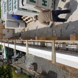 firma budowlana Kania - Budowa ściany z białych bloczków betonowych, zbrojonej metalowymi prętami i drewnianym szalunkiem. Widoczny pracownik w kasku i zapas bloczków na paletach oraz betoniarka w tle.