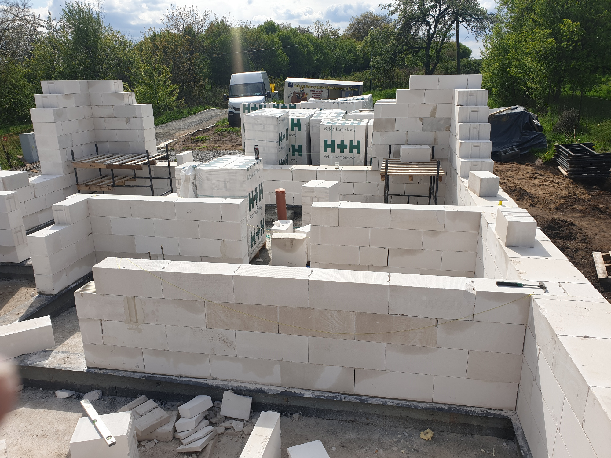 Wznoszenie ścian parteru z bloczków betonu komórkowego H+H na fundamencie, widoczne palety z materiałem, w tle zaparkowany bus firmy budowlanej.