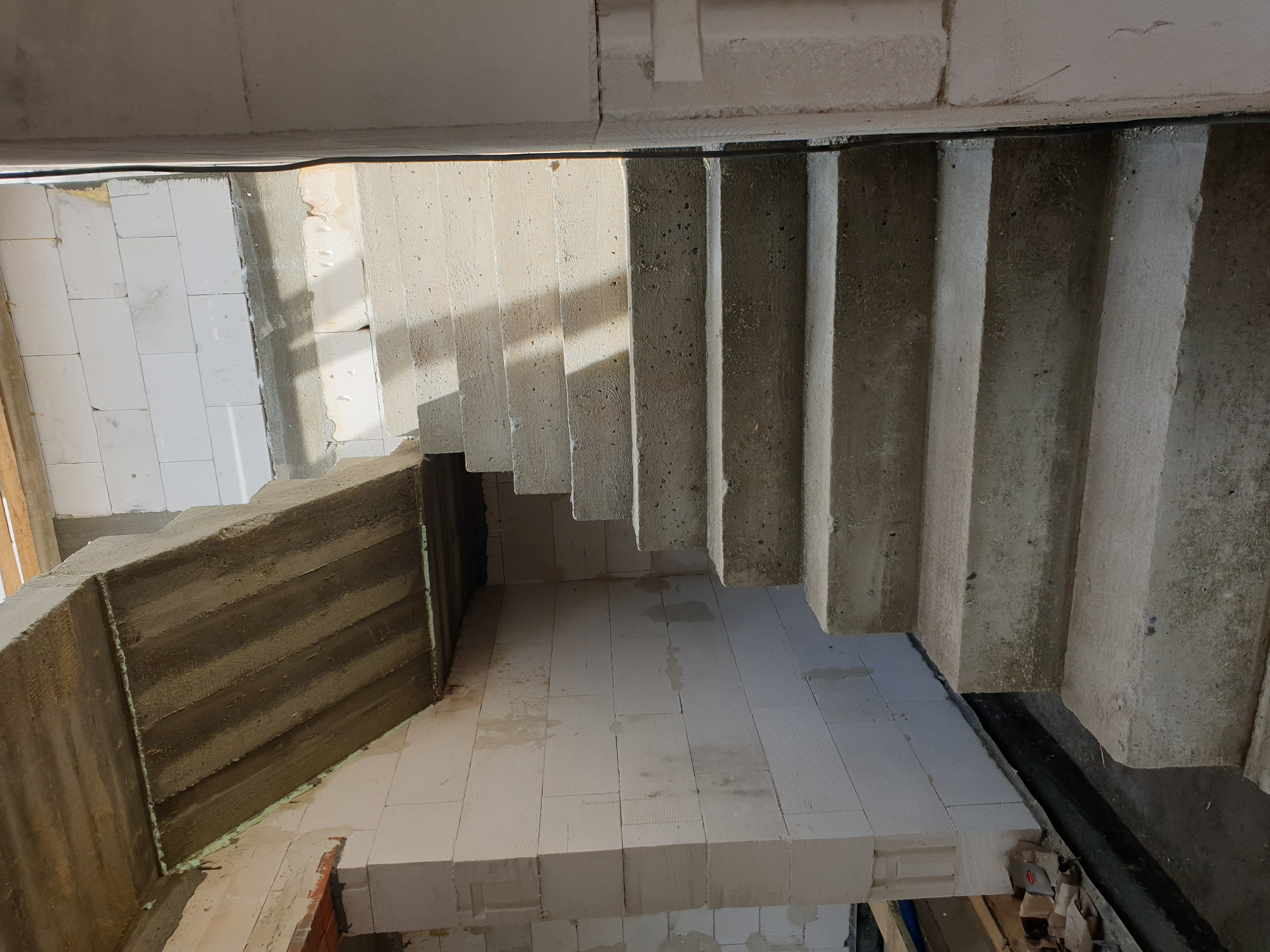 Wnętrze w trakcie budowy z widocznymi surowymi schodami betonowymi, ścianami z bloczków i częściowo otynkowaną ścianą boczną.