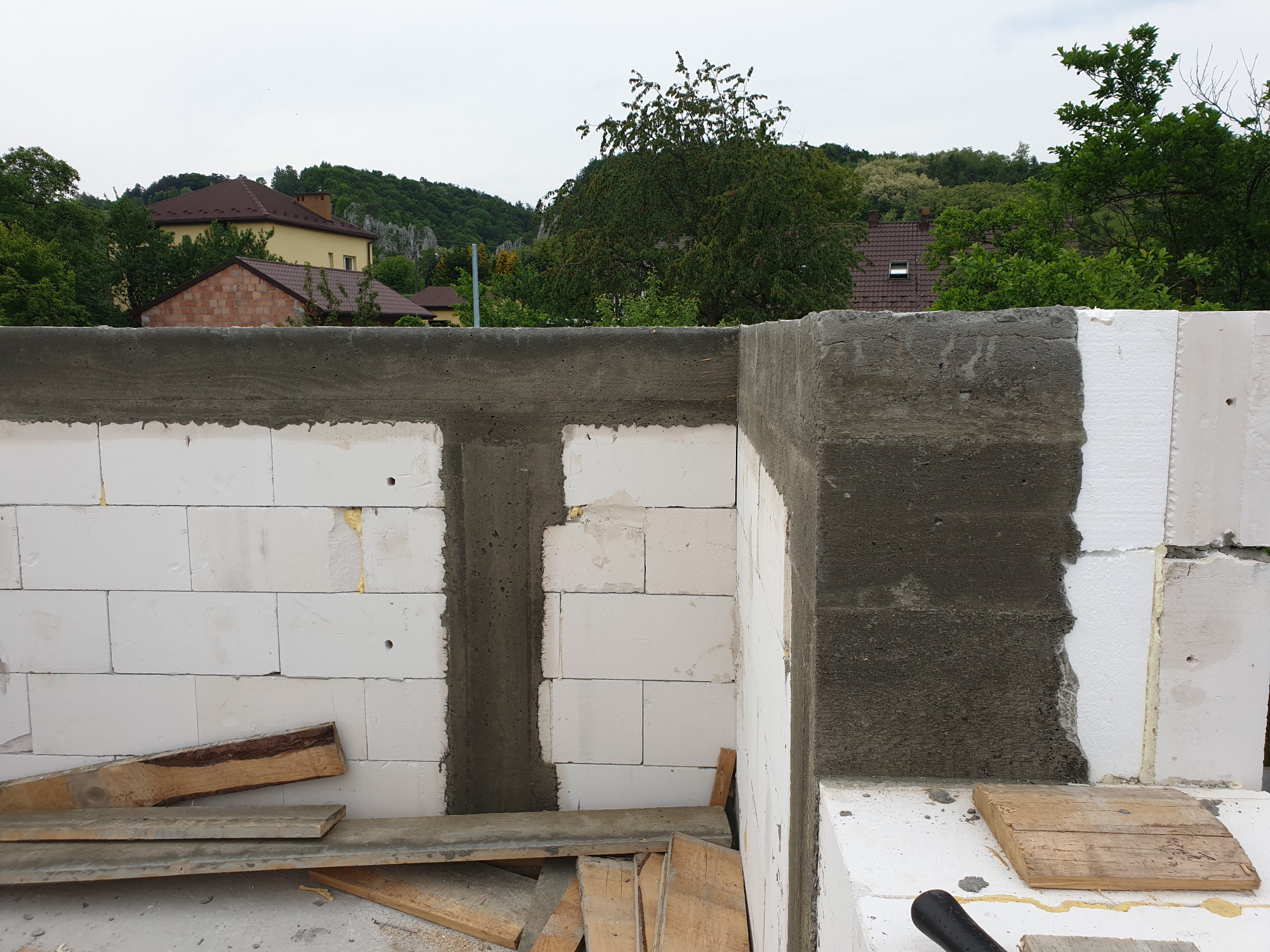 Wznoszenie ścian domu murowanego z betonu komórkowego, z widocznym zbrojonym narożnikiem i fragmentami izolacji termicznej, w tle zabudowa jednorodzinna i zieleń.