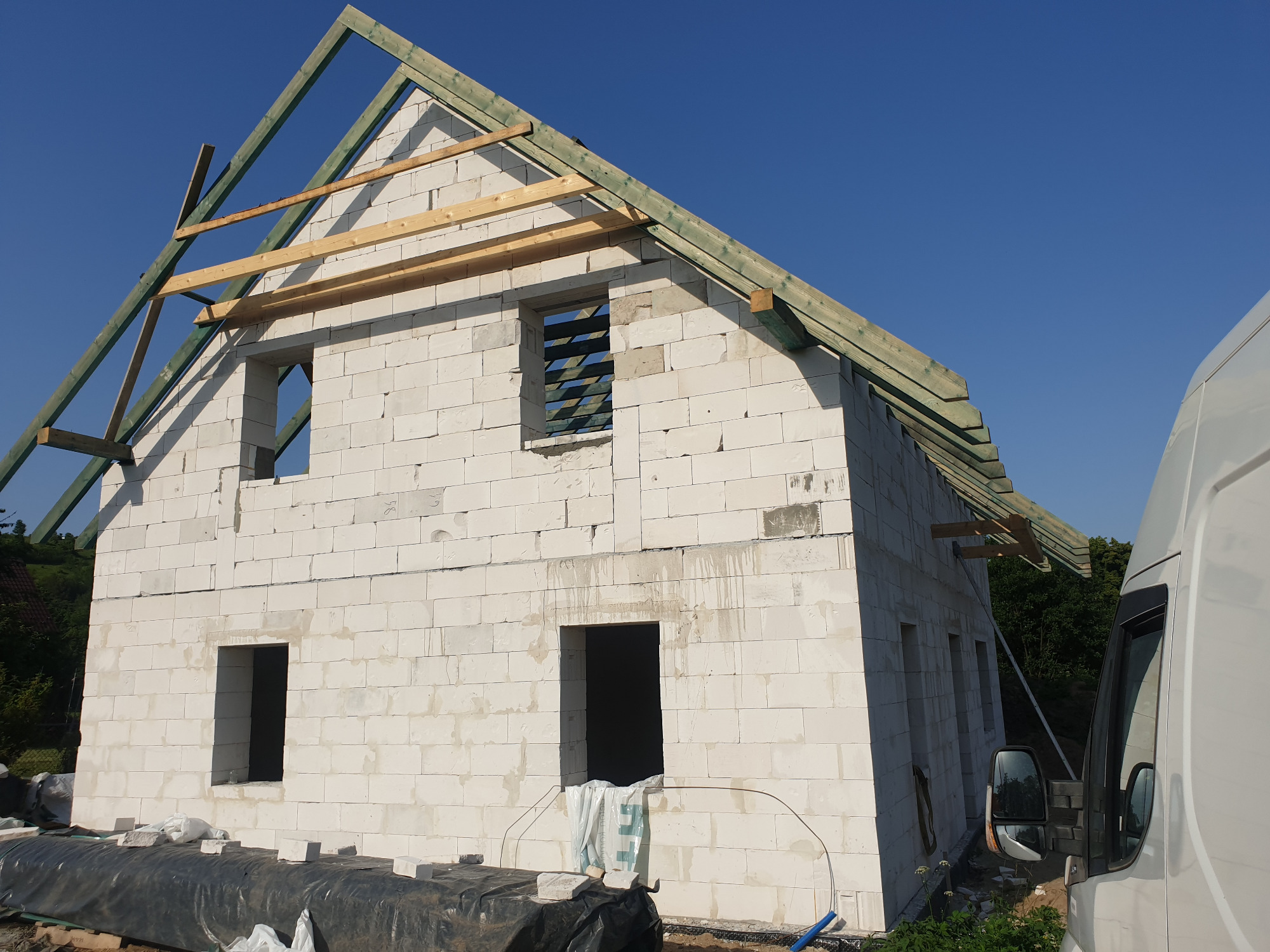 Surowy stan domu z białych bloczków, widoczne otwory okienne i drzwiowe, konstrukcja dachu z zielonych łat i drewnianych belek, częściowo zakryty biały samochód dostawczy po prawej stronie.