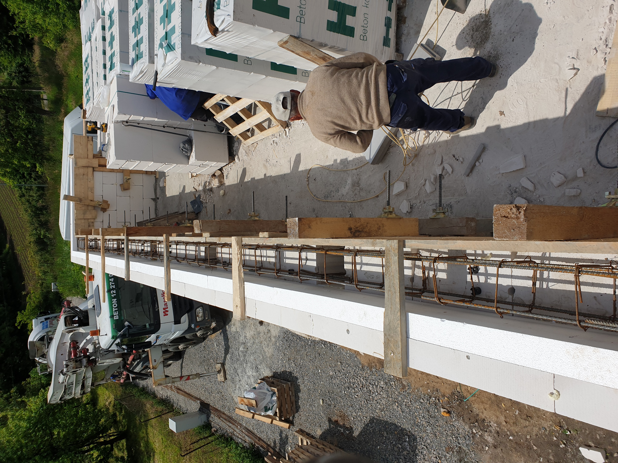 Budowa ściany z białych bloczków betonowych, zbrojonej metalowymi prętami i drewnianym szalunkiem. Widoczny pracownik w kasku i zapas bloczków na paletach oraz betoniarka w tle.