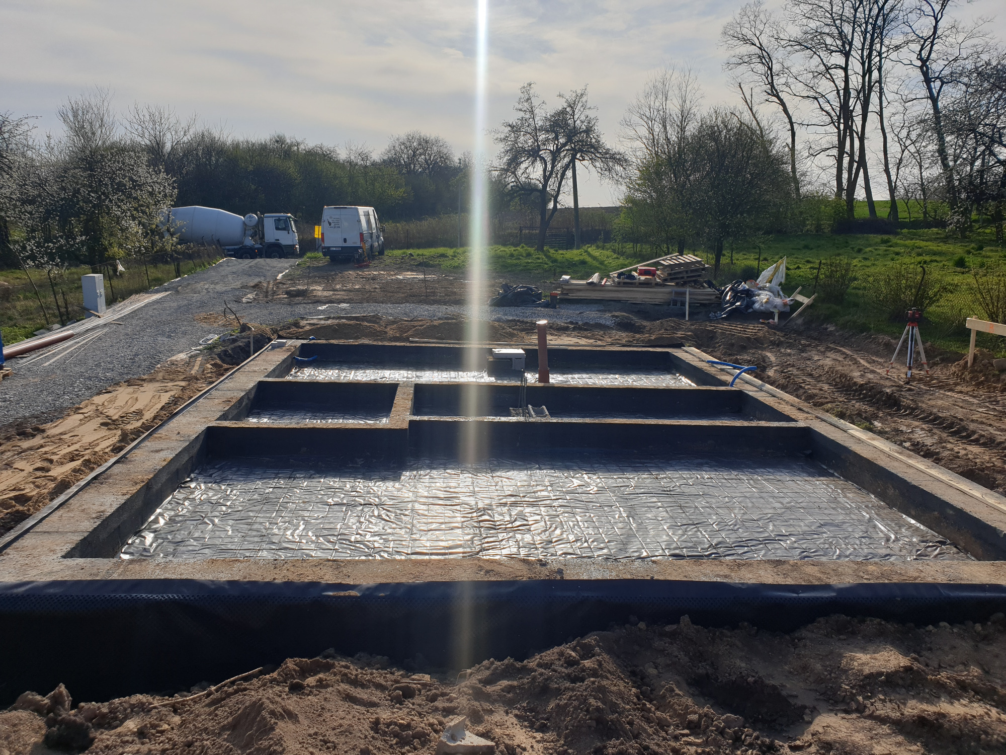 Zalane betonem szalunki fundamentowe z hydroizolacją, widoczne przygotowane podłoże, na dalszym planie betoniarka i samochód dostawczy.