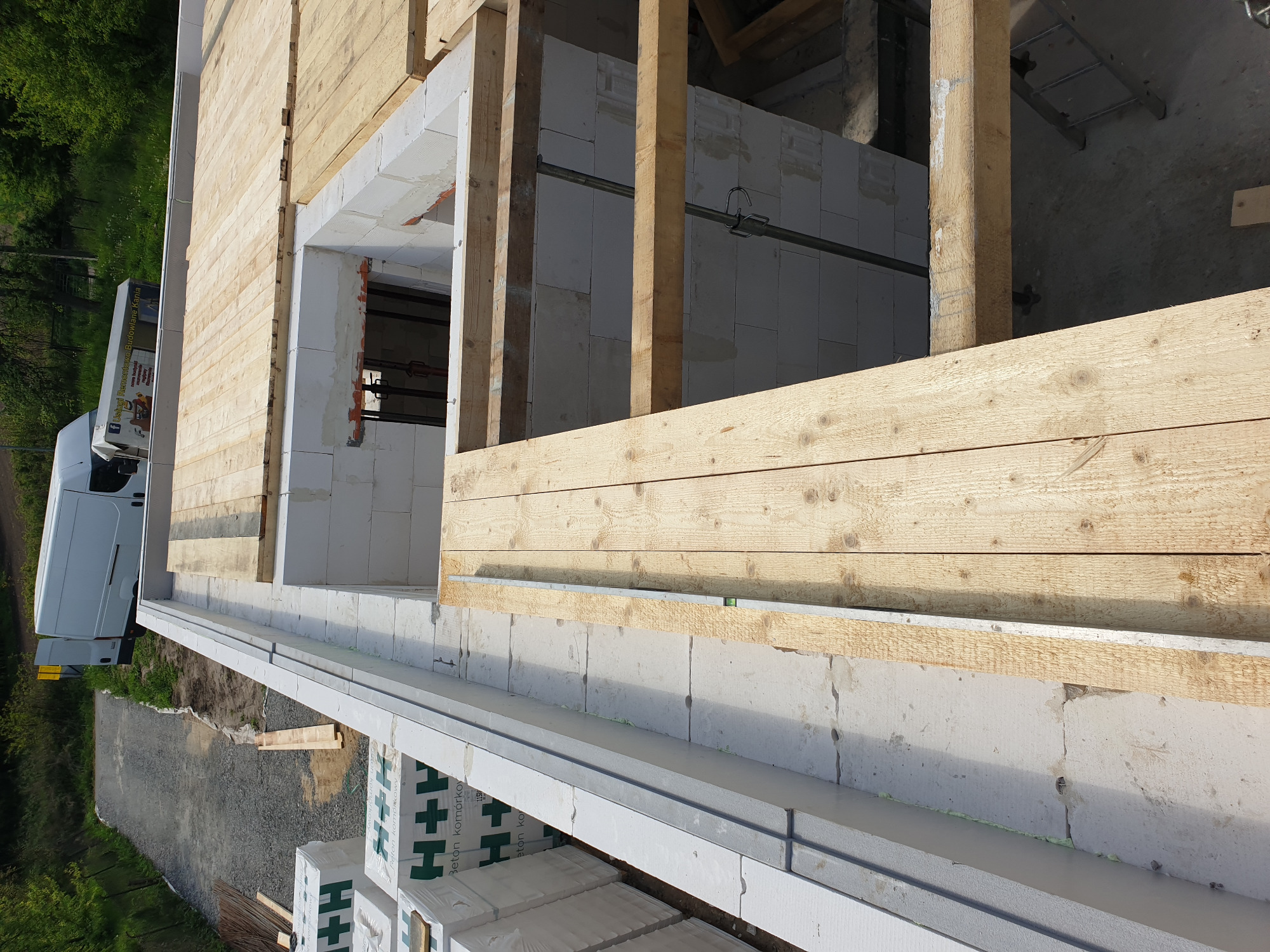 Wznoszenie ścian parteru z betonu komórkowego, widoczne drewniane stemple podtrzymujące konstrukcję, na zewnątrz zaparkowany biały bus firmy budowlanej.