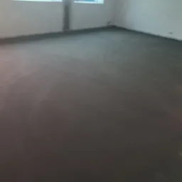 Świeżo wylana, szara posadzka jastrychowa w surowym pomieszczeniu z dwoma oknami. Widoczne ślady po instalacjach na ścianie.