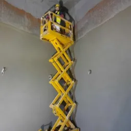 Pracownik w ż&oacute;łtym kasku i kamizelce na podnośniku nożycowym, wykonujący prace wykończeniowe na wysokości, widoczny fragment surowego sufitu i świeżo nałożona gładź na ścianach.