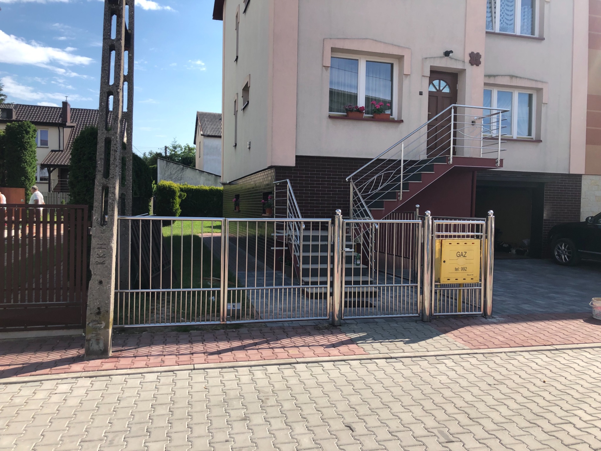 Nierdzewna brama i balustrada schodowa przed domem z żółtą skrzynką gazową na słupku bramy.
