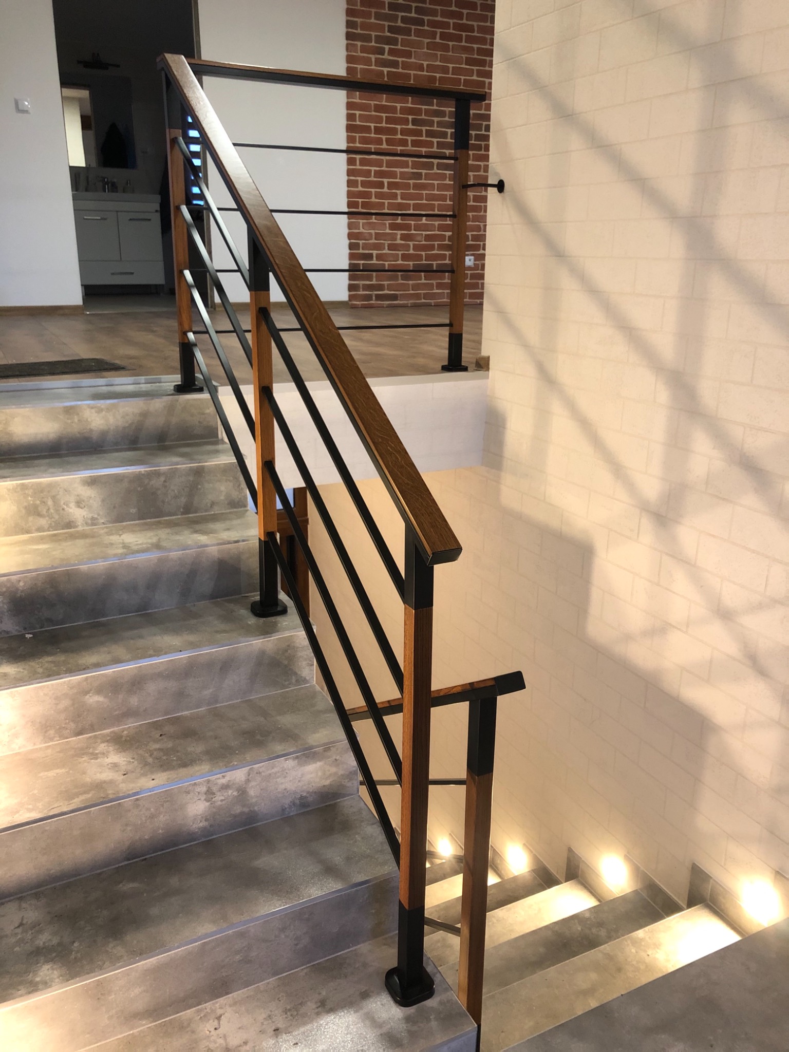 Schody z drewnianą poręczą i czarnymi, metalowymi prętami, oświetlone punktowo wzdłuż ściany z cegieł w tle.