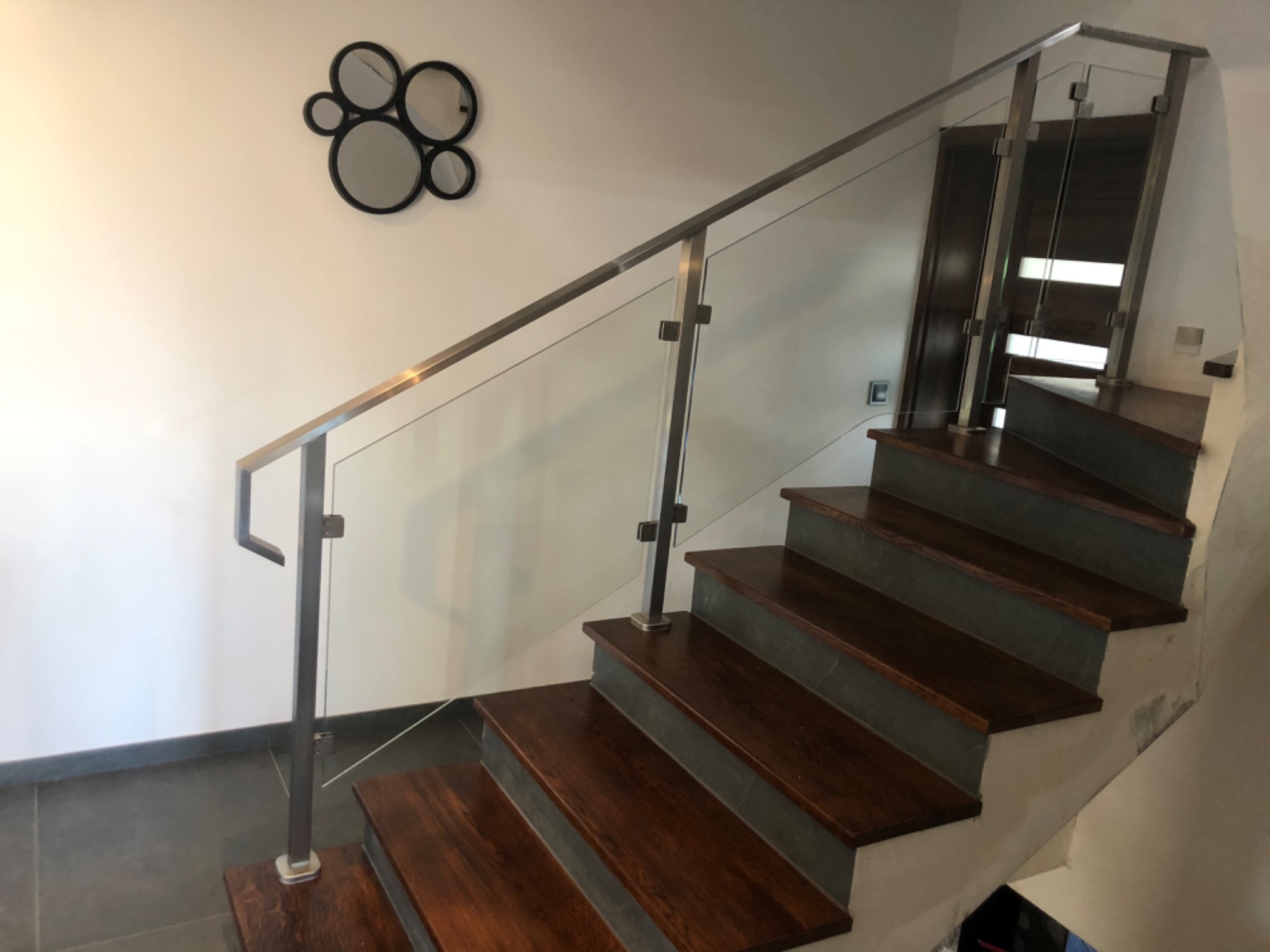 Wewnętrzne schody z drewnianymi stopniami i szklaną balustradą ze stalową ramą, na białej ścianie dekoracja z luster.