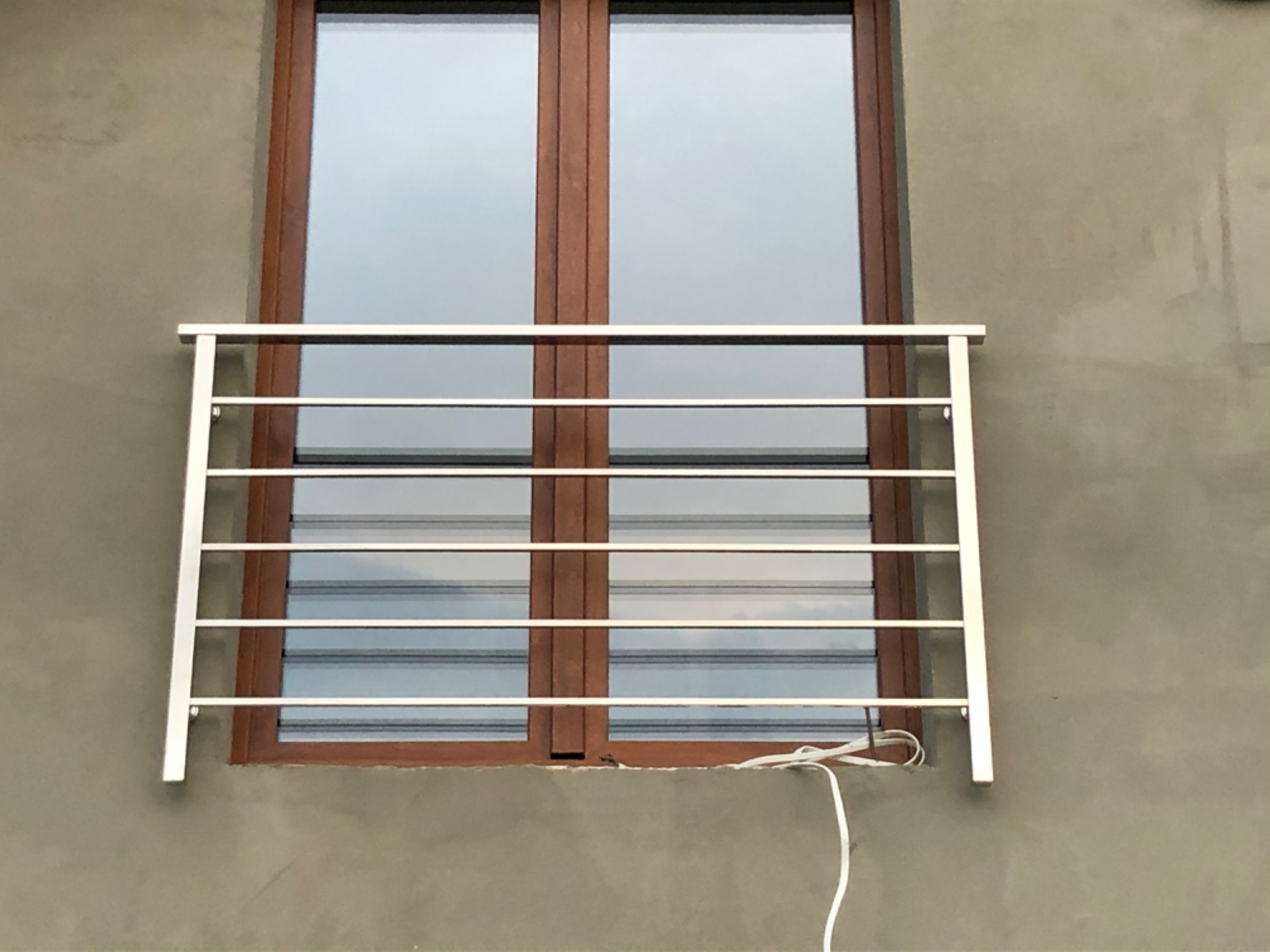 Nierdzewna balustrada okienna z poziomymi szczeblami, zamontowana na szorstkiej, szarej elewacji budynku, widok z zewnątrz, kabel elektryczny zwisający wzdłuż ściany.