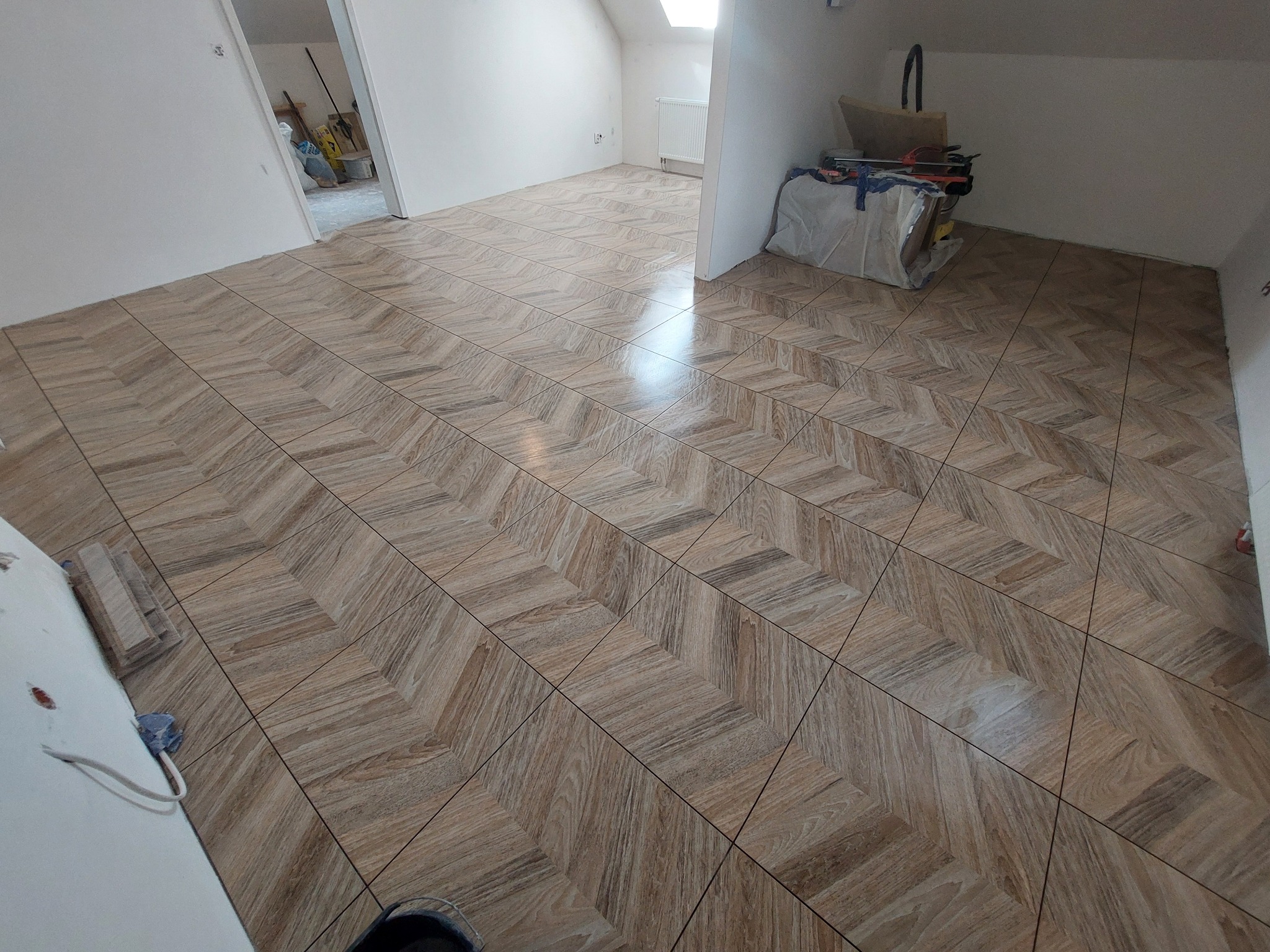 Wykończona podłoga w jodełkę z płytek imitujących drewno w jasnym pomieszczeniu na poddaszu, narzędzia glazurnicze w tle.