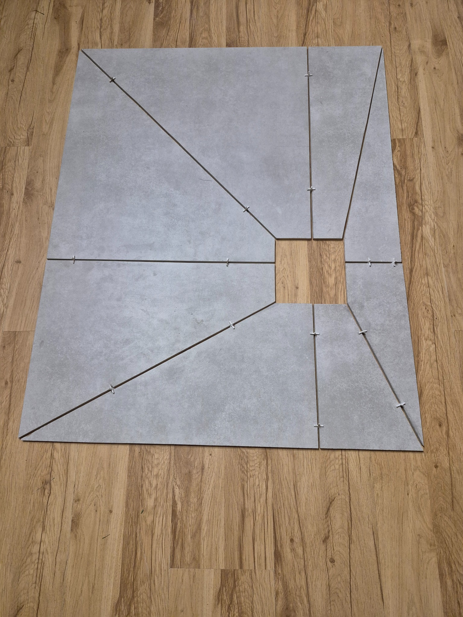 Układanie szarych płytek na podłodze z drewnopodobnych paneli. Płytki ułożone w kwadrat z centralnym otworem, z użyciem krzyżyków dystansowych. Widok z góry.