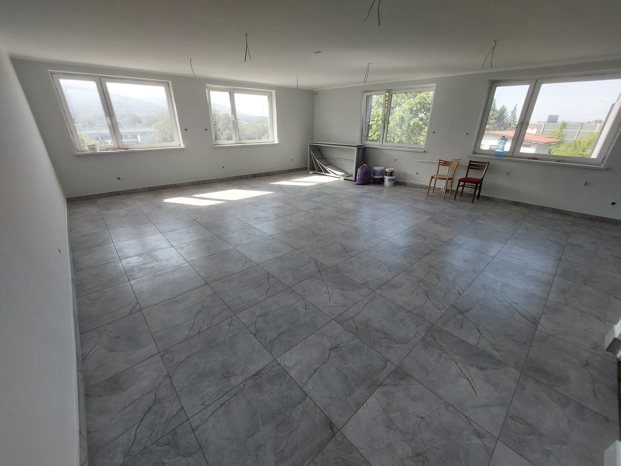 Wykończone wnętrze: szare, kwadratowe płytki podłogowe z wzorem marmuru, białe ściany, duże okna z widokiem na zieleń. Dwa krzesła i puszki farby w tle.