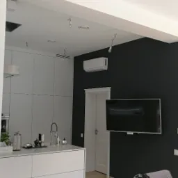 Wnętrze rezydencji z zamontowaną jednostką klimatyzacyjną na ścianie, widoczne elementy kuchni i salonu w nowoczesnym stylu, z elementami w trakcie wykańczania.