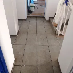 Ułożona szara terakota podłogowa w korytarzu, widoczne białe ściany, zabezpieczone schody i wiadro farby w tle.