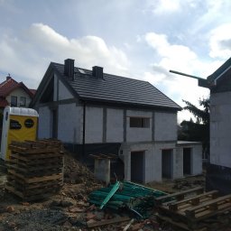 DEC-BUD - Niedokończony dom z szarego bloczka, z czarnym dachem, widoczny z zewnątrz. Na pierwszym planie palety i gruz, w tle błękitne niebo z chmurami. Widoczne elementy instalacji.