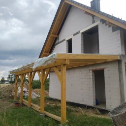 DEC-BUD - Budowa domu z białego bloczka, widoczna drewniana konstrukcja zadaszenia tarasu na etapie wznoszenia. Niebo pochmurne.