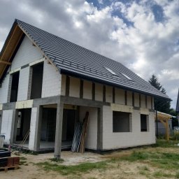 DEC-BUD - Surowy dom jednorodzinny z szarej cegły silikatowej pod ciemnym dachem z dachówki ceramicznej. Widoczne okna dachowe i brak okien w ścianach.