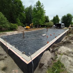 DEC-BUD - Przygotowany fundament pod budowę domu z hydroizolacją, zbrojeniem i częściowo wylaną ławą. Na działce stoi koparka i bus.