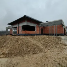 Surowy stan domu z czerwonej cegły i betonu pod pochmurnym niebem, widok z poziomu gruntu na nierówny teren budowy.