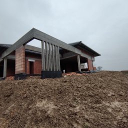 DEC-BUD - Surowy stan domu w trakcie budowy: widoczne ceglane ściany, betonowe słupy i fundamenty na tle ziemi. Nisko zawieszone, pochmurne niebo.