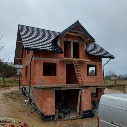 DEC-BUD - Dwukondygnacyjny dom w trakcie budowy, z czarną dachówką i widocznymi ceglanymi ścianami. Na pierwszym planie gruz budowlany i częściowo widoczny samochód dostawczy.