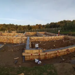 Szeroki kadr na drewniany szalunek fundamentów domu jednorodzinnego w trakcie budowy, z widocznymi zbrojeniami i częściowo wylanym betonem, tło: drzewa i błękitne niebo.