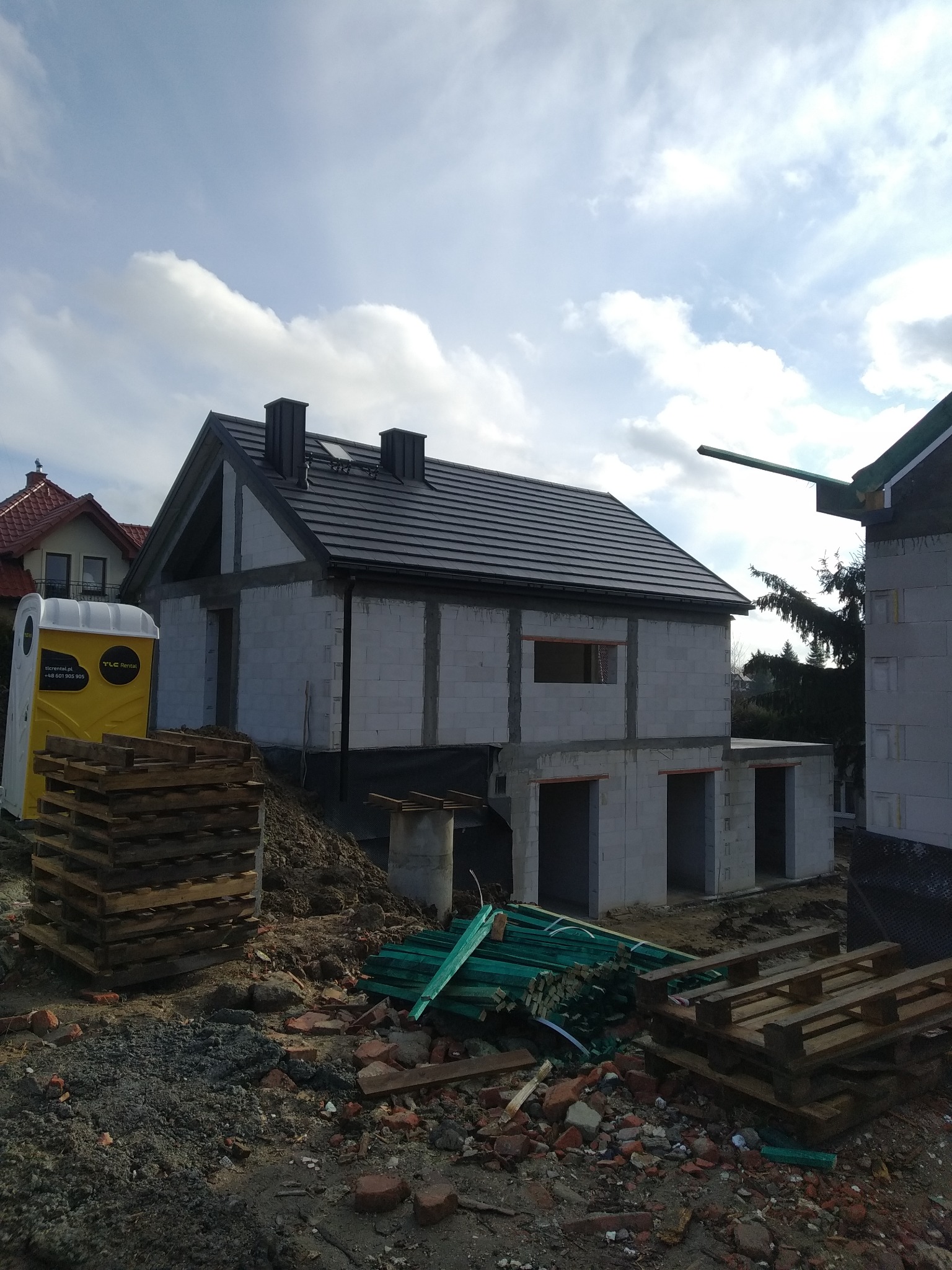 Niedokończony dom z szarego bloczka, z czarnym dachem, widoczny z zewnątrz. Na pierwszym planie palety i gruz, w tle błękitne niebo z chmurami. Widoczne elementy instalacji.