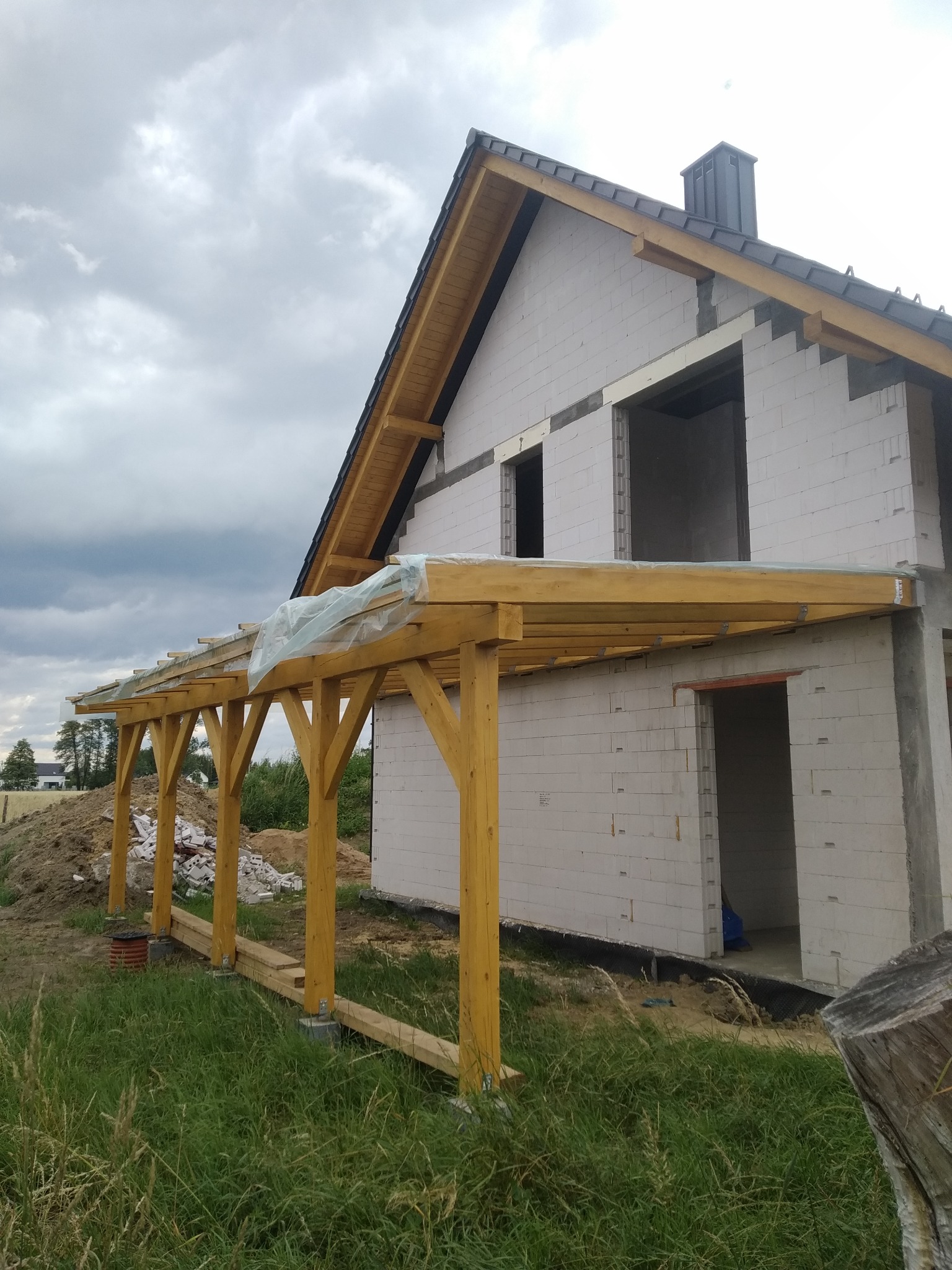Budowa domu z białego bloczka, widoczna drewniana konstrukcja zadaszenia tarasu na etapie wznoszenia. Niebo pochmurne.