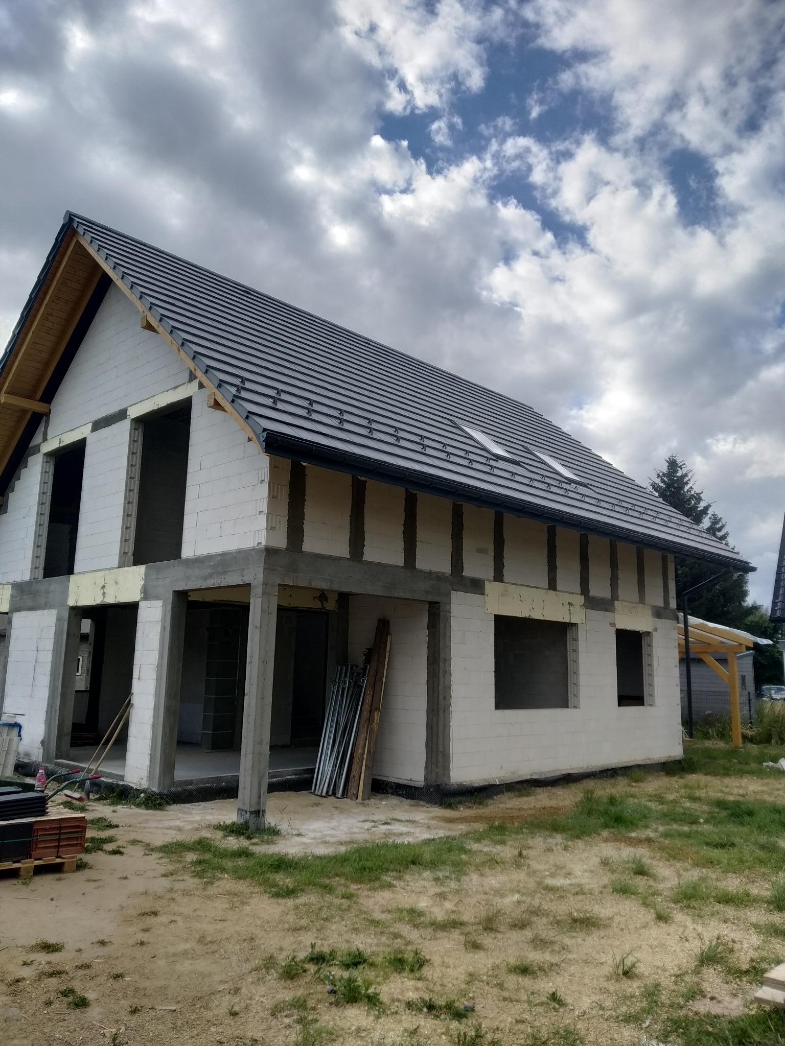 Surowy dom jednorodzinny z szarej cegły silikatowej pod ciemnym dachem z dachówki ceramicznej. Widoczne okna dachowe i brak okien w ścianach.