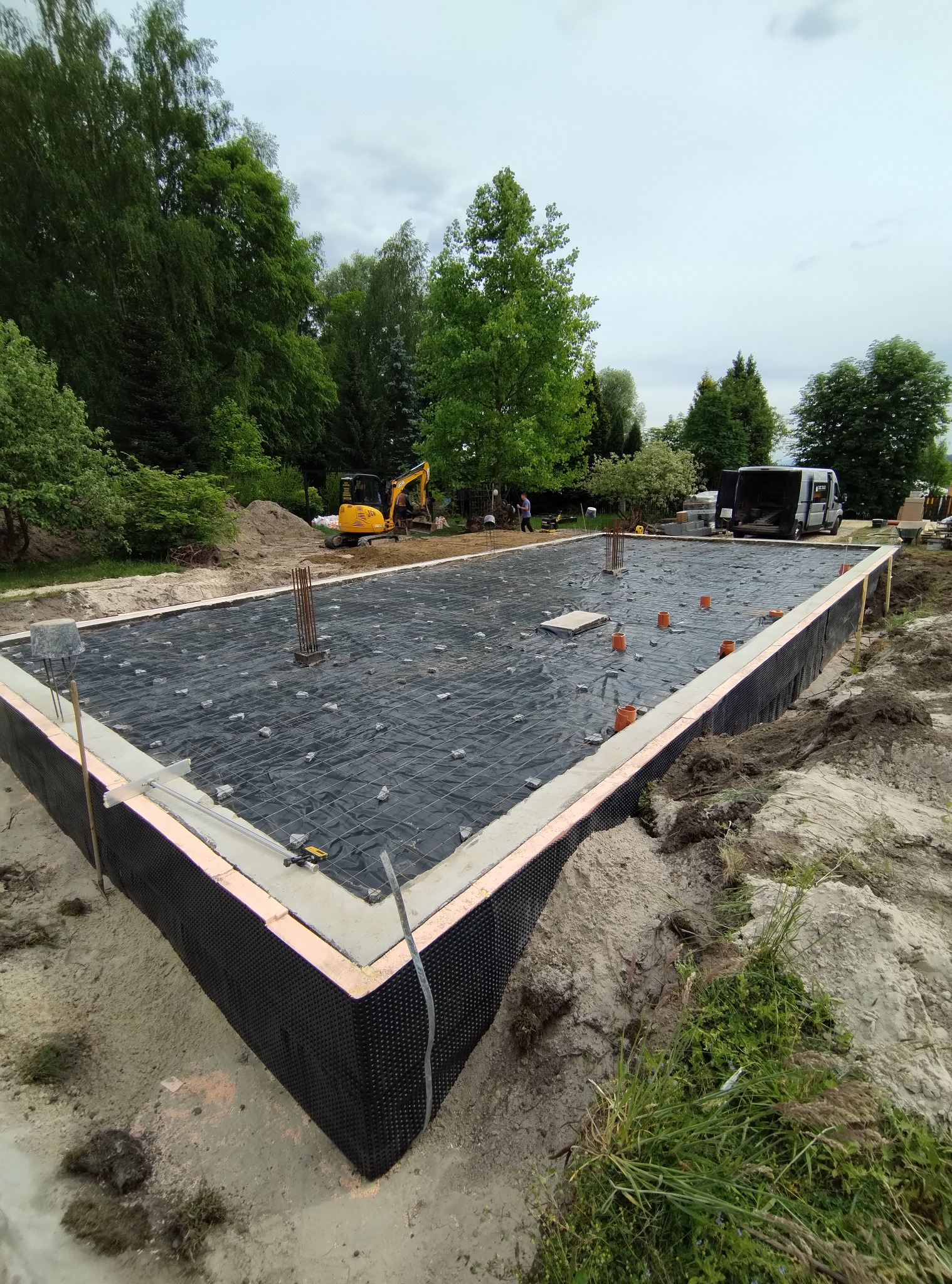 Przygotowany fundament pod budowę domu z hydroizolacją, zbrojeniem i częściowo wylaną ławą. Na działce stoi koparka i bus.