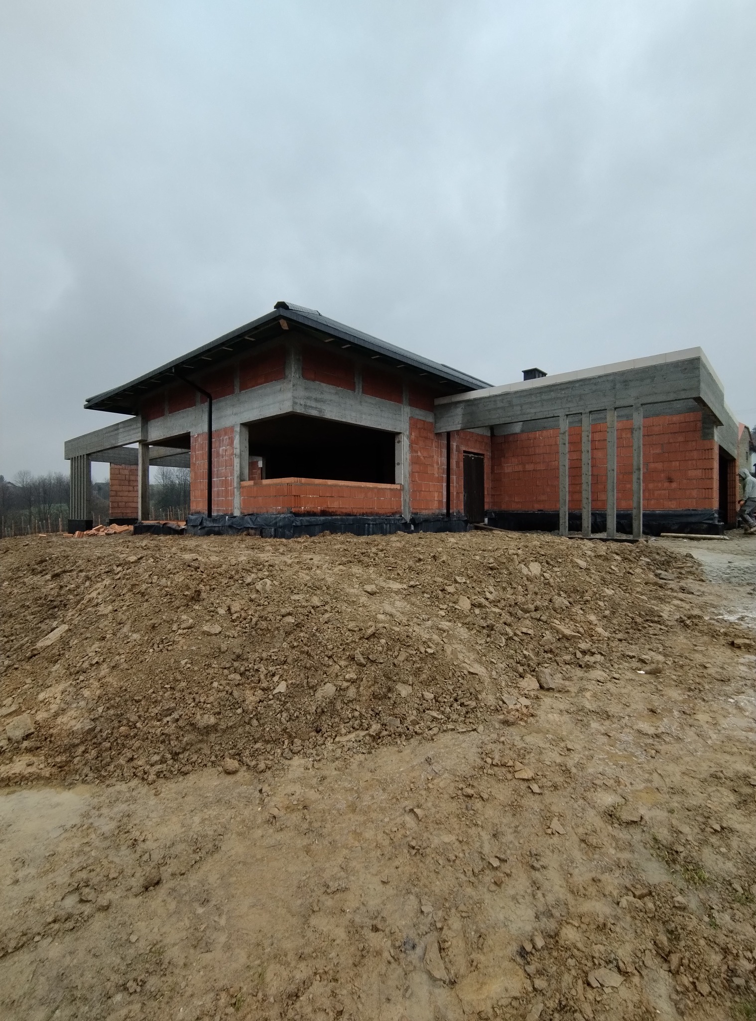 Surowy stan domu z czerwonej cegły i betonu pod pochmurnym niebem, widok z poziomu gruntu na nierówny teren budowy.