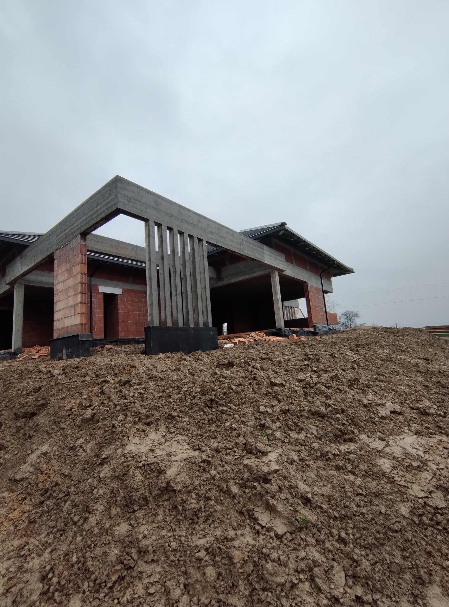 Surowy stan domu w trakcie budowy: widoczne ceglane ściany, betonowe słupy i fundamenty na tle ziemi. Nisko zawieszone, pochmurne niebo.