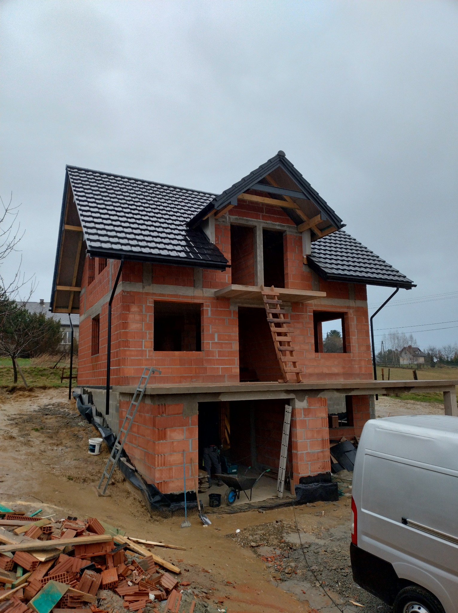 Dwukondygnacyjny dom w trakcie budowy, z czarną dachówką i widocznymi ceglanymi ścianami. Na pierwszym planie gruz budowlany i częściowo widoczny samochód dostawczy.
