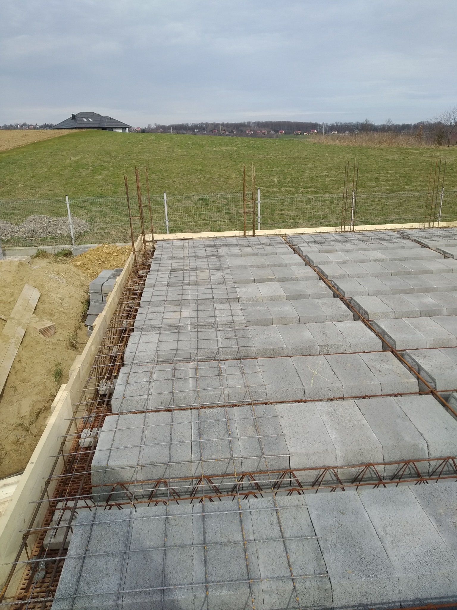 Zbrojenie i szalunek pod wylewkę fundamentów domu. Widoczne bloczki betonowe, stalowe pręty i drewniany szalunek. W tle zielone pole i dom.