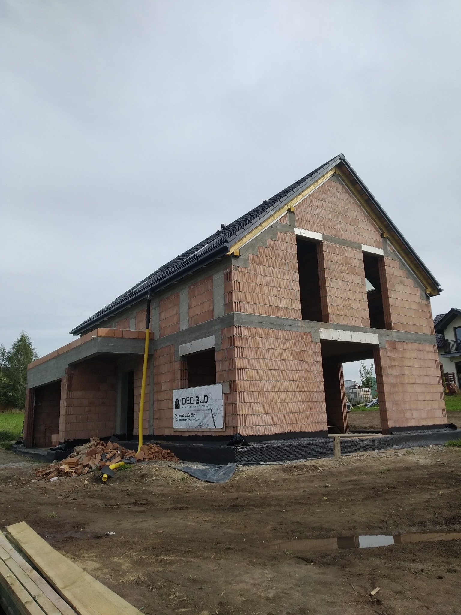 Surowy, dwukondygnacyjny dom z czerwonej cegły w trakcie budowy. Widoczne niezadaszone wejście, brak okien i drzwi, dach z czarną dachówką. Teren wokół w stanie surowym.