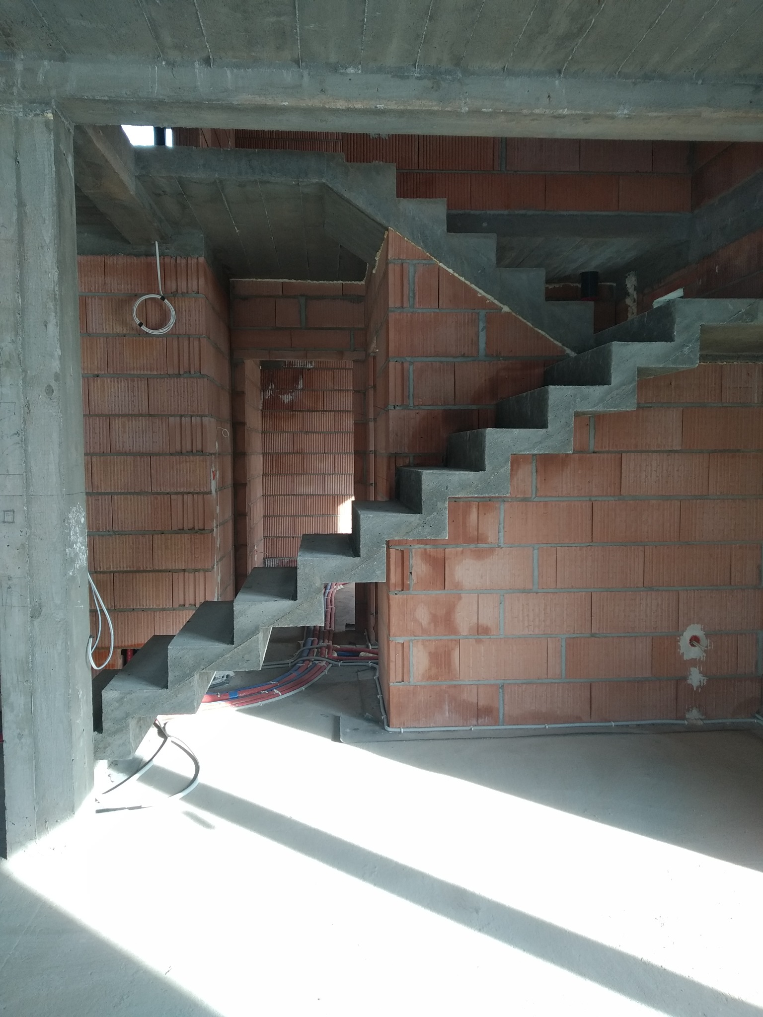 Surowe schody betonowe w trakcie budowy, osadzone w murze z czerwonej cegły. Widoczne instalacje i surowy betonowy strop. Wpadające światło słoneczne.