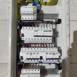 Rozdzielnica elektryczna w trakcie montażu, widoczne bezpieczniki Schneider i Hager z ręcznie napisanymi oznaczeniami obwodów, kable w listwach zaciskowych, surowa ściana z betonu komórkowego w tle.