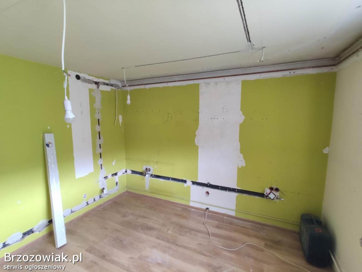 Wnętrze w trakcie remontu z widocznymi przewodami elektrycznymi w ścianach, częściowo zakryte gipsem, poziomica oparta o ścianę, surowa żarówka zwisająca z sufitu.