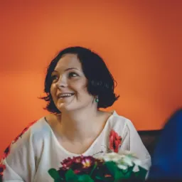 Uśmiechnięta kobieta z krótkimi, ciemnymi włosami, w bluzce z kwiatowym wzorem, siedząca przy stole z bukietem kwiatów na tle pomarańczowej ściany.