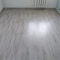 Jasne panele podłogowe z widoczną strukturą drewna, ułożone w pustym pokoju z białymi ścianami i grzejnikiem.