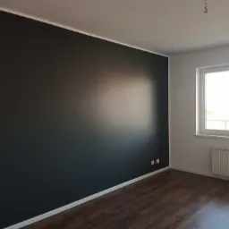 Pomieszczenie po odświeżeniu: ciemnoszara ściana kontrastuje z białym sufitem i listwami, brązowe panele podłogowe, okno z widokiem na zewnątrz, biały grzejnik pod oknem, gniazdka elektryczne przy...