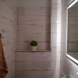 Wykończona łazienka z białymi płytkami imitującymi drewno, podświetlanym lustrem, ręcznikiem na haczyku i rośliną doniczkową na półce.